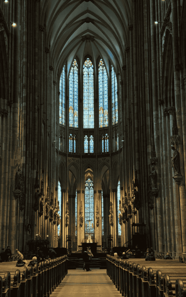 The grand interior of Cologne Cathedral features soaring vaulted ceilings, intricate stone columns, and tall stained-glass windows behind the altar.