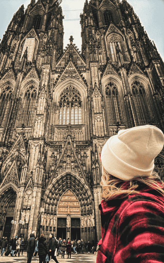 Kate, wearing a white beanie and red plaid jacket, looks up at the towering gothic facade of Cologne Cathedral.