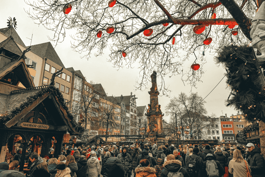 Crowds browse the bustling Cologne Christmas Market, framed by red hanging lanterns and festive stalls around a tall decorative monument.