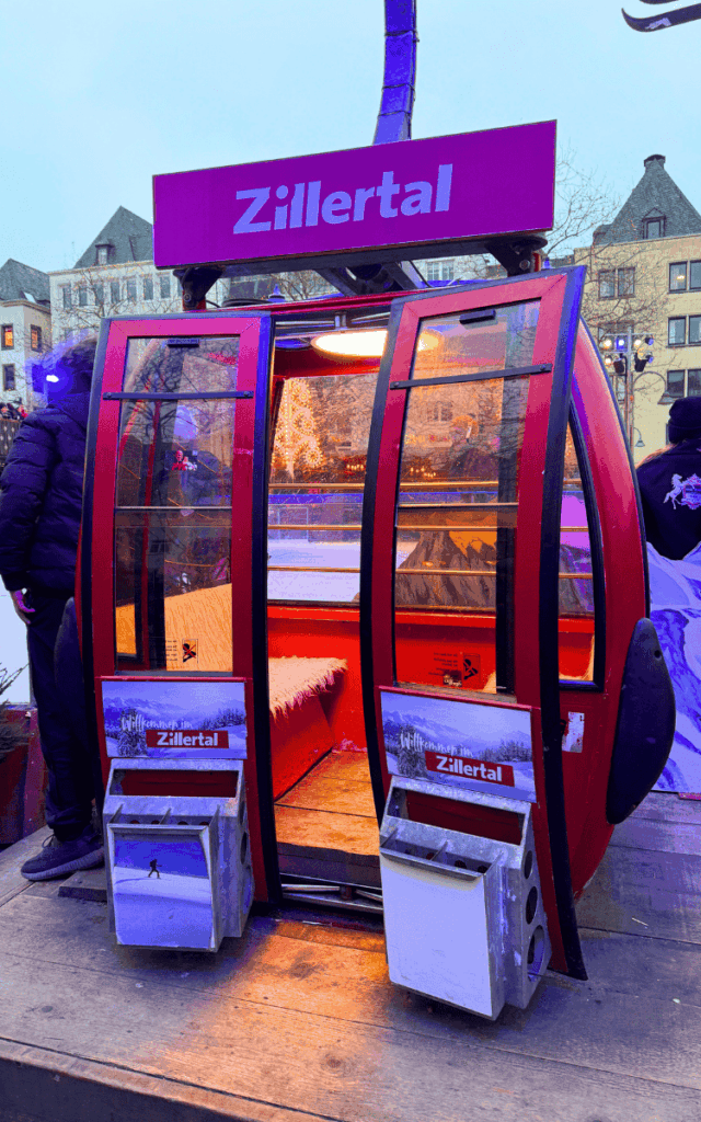 A red ski gondola cabin labeled “Zillertal” sits open as part of a festive display at the Cologne Christmas Market.