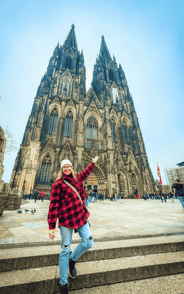 Kate, smiling in a red plaid jacket and white beanie, stands on the steps and points up at the towering Cologne Cathedral.