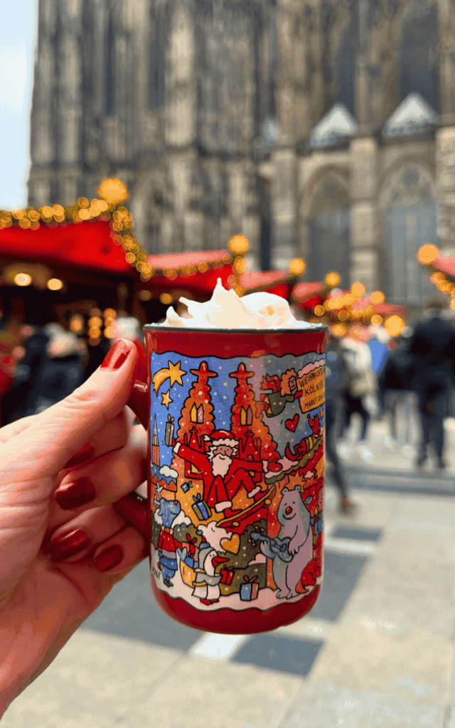 A hand with red-painted nails holds a festive Christmas mug of whipped cream-topped hot chocolate in front of Cologne Cathedral at the Christmas Market.