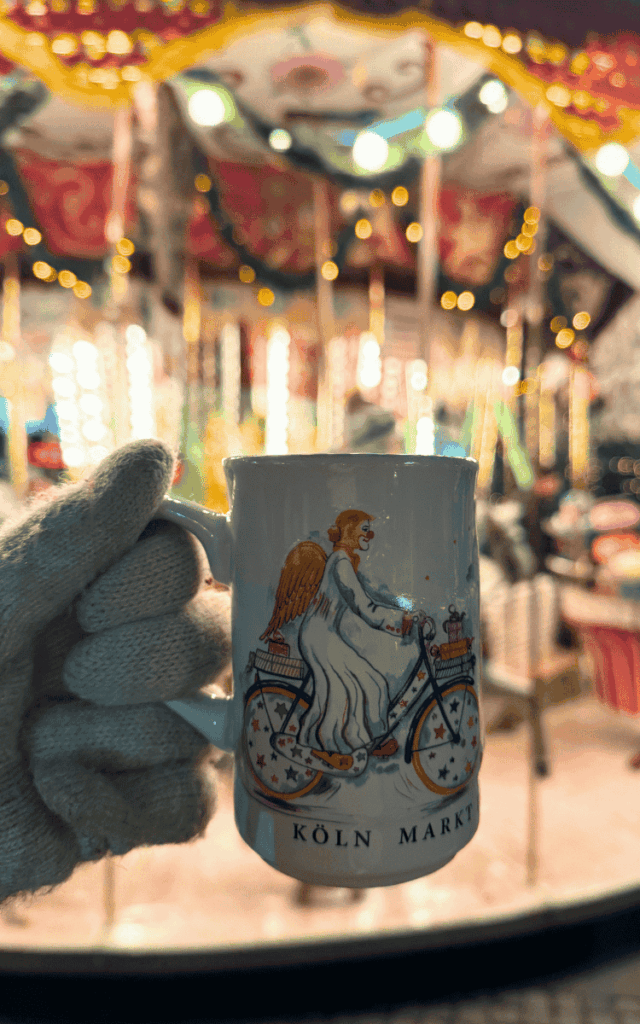 A gloved hand holds a white “Köln Markt” mug featuring an angel riding a bicycle in front of a glowing carousel.