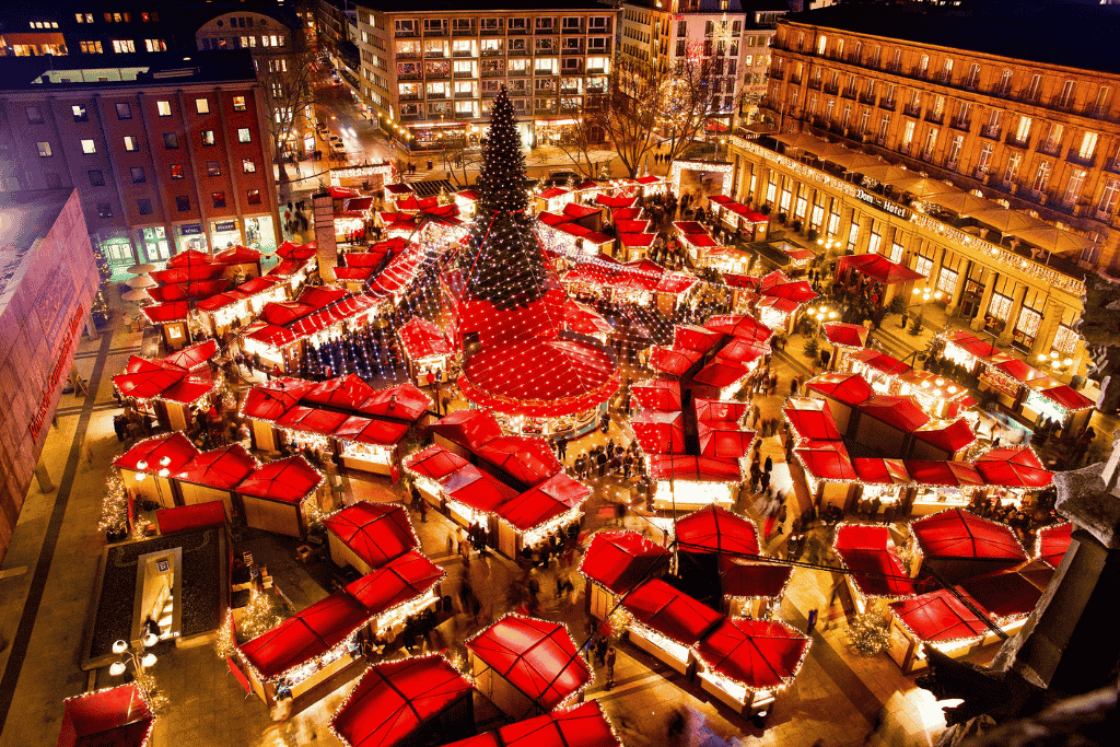 An aerial view of the Cologne Cathedral Christmas Market shows rows of red-roofed stalls radiating from a towering, light-covered Christmas tree at night.