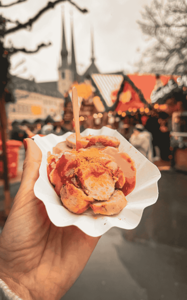 Kate holds a paper tray of currywurst topped with ketchup and curry powder, with the festive Luxembourg Christmas market and cathedral spires blurred in the background.
