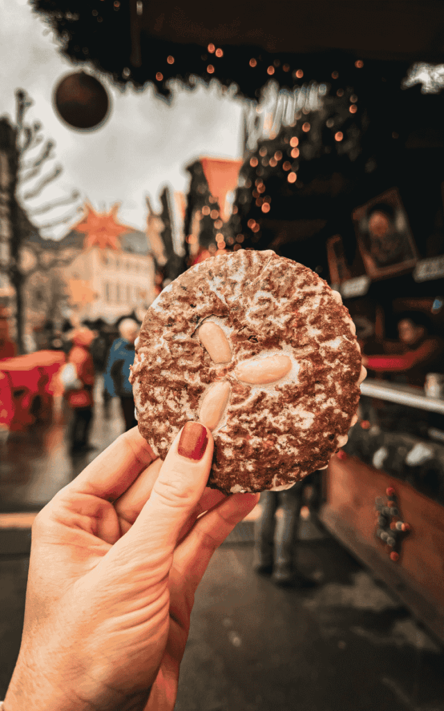 Kate displays a round, powdered gingerbread cookie topped with whole almonds at the Luxembourg Christmas market, with twinkling lights and a wooden stall behind her.