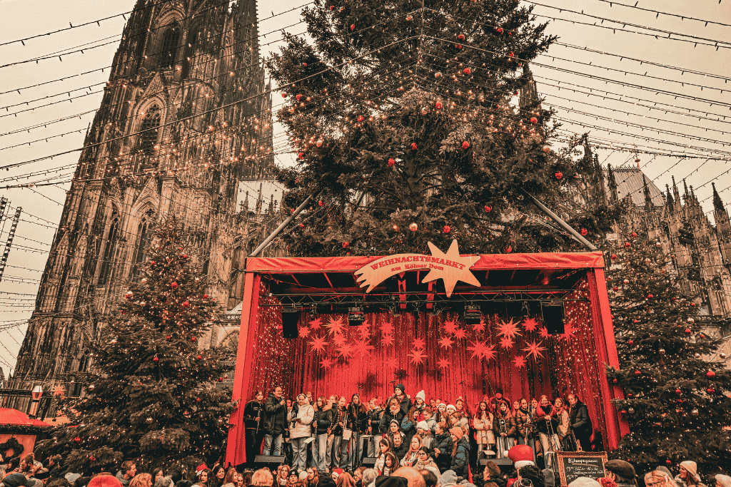 A festive choir performs on a red-lit stage beneath a towering Christmas tree at the Cologne Cathedral Christmas Market.