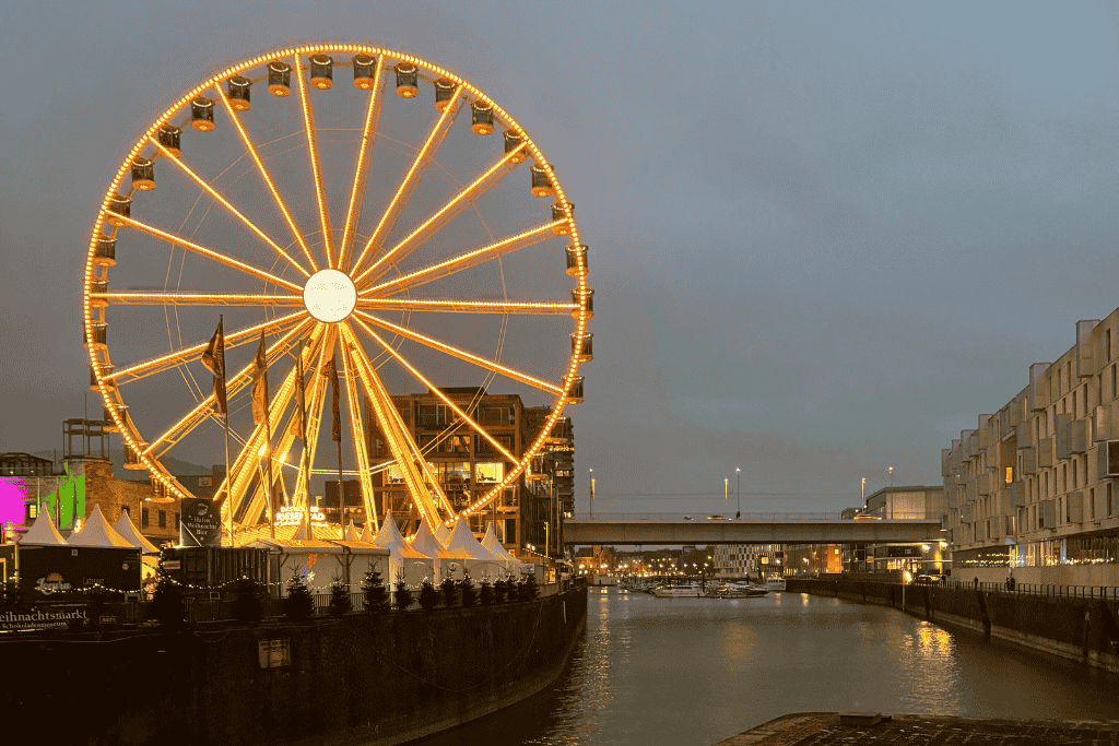 A brightly lit Ferris wheel glows against the evening sky beside the waterfront at the Cologne Christmas Market.