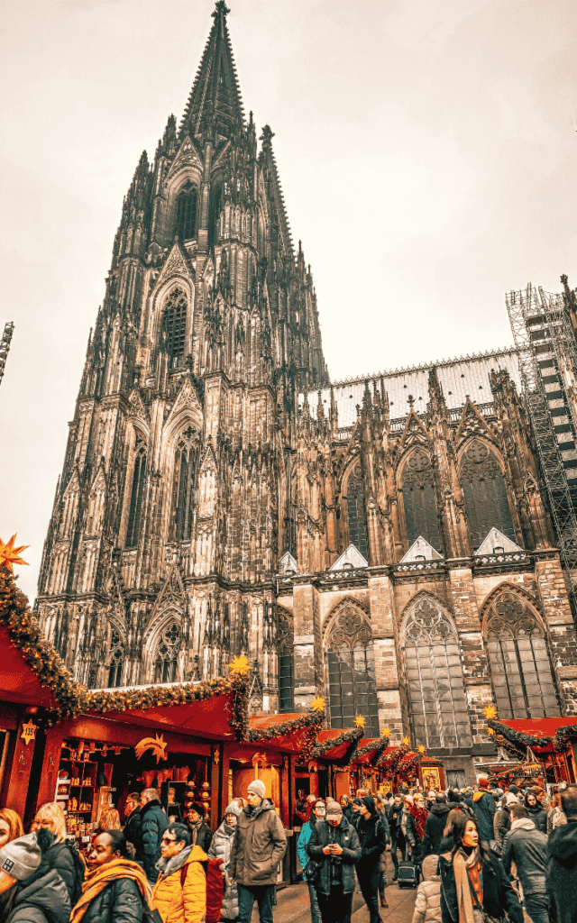 Visitors browse red-roofed, garland-trimmed stalls at the bustling Cologne Cathedral Christmas Market.