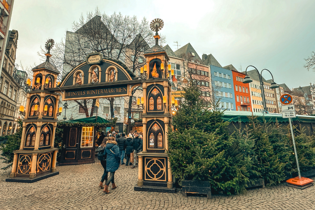 Crowds gather under the ornate wooden archway of Heinzel’s Wintermärchen at the Cologne Christmas Market, surrounded by twinkling lights and festive evergreen trees.