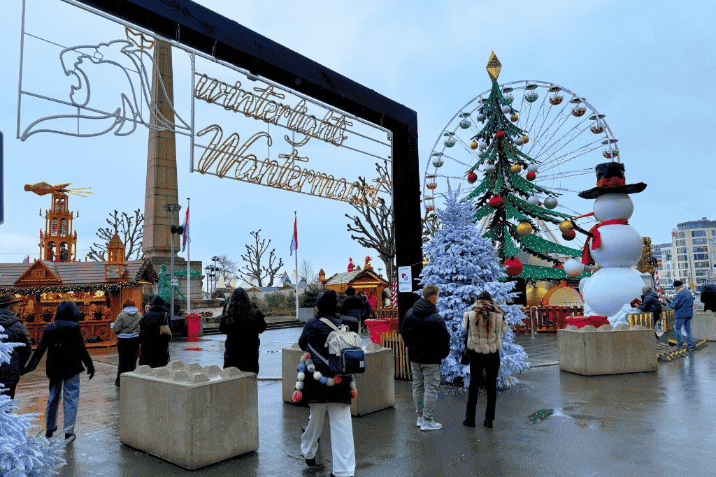 Kate stands beneath the "Winterlights Wantermaart" sign at the Luxembourg Christmas market, facing a festive scene with a giant Ferris wheel, oversized Christmas tree, and inflatable snowman.