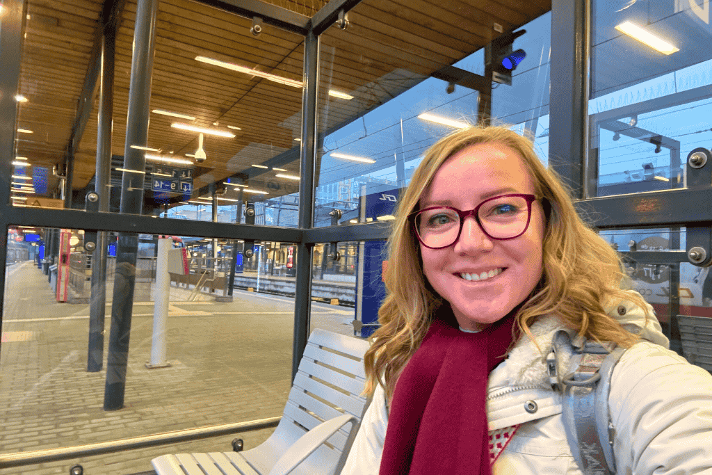 Kate smiles while sitting on a bench at a modern train station, with glass walls, wooden ceilings, and train platforms visible in the background.