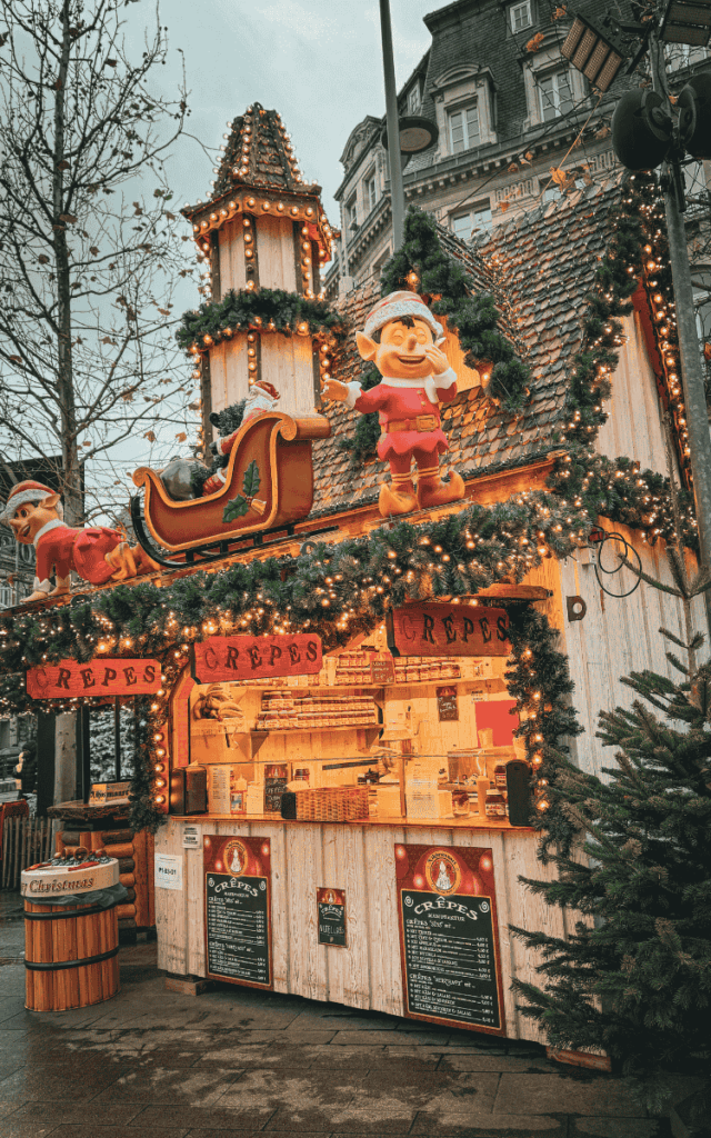 A brightly lit Christmas market crêpe stall decorated with garlands, Santa’s sleigh, and cheerful elf figures glows warmly against the evening sky