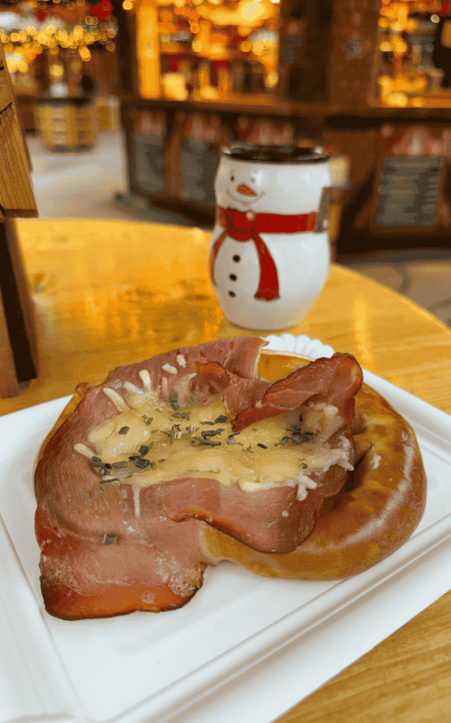 A festive market table holds a soft pretzel topped with melted cheese and ham, with a snowman-shaped mug of hot drink in the background.