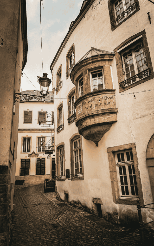 A narrow cobblestone street in Luxembourg City winds past historic buildings, including one with a stone bay window inscribed with “Mir wölle bleiwe wat mir sin.”