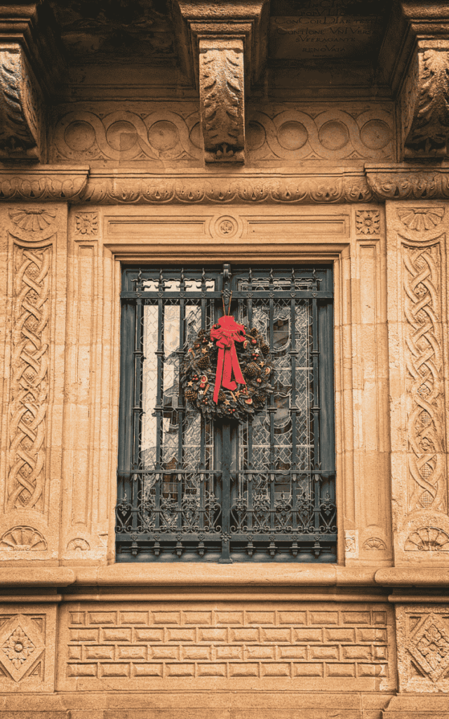 A black wrought-iron window is decorated with a festive Christmas wreath adorned with dried fruit and a bright red bow, set in an ornate stone facade.