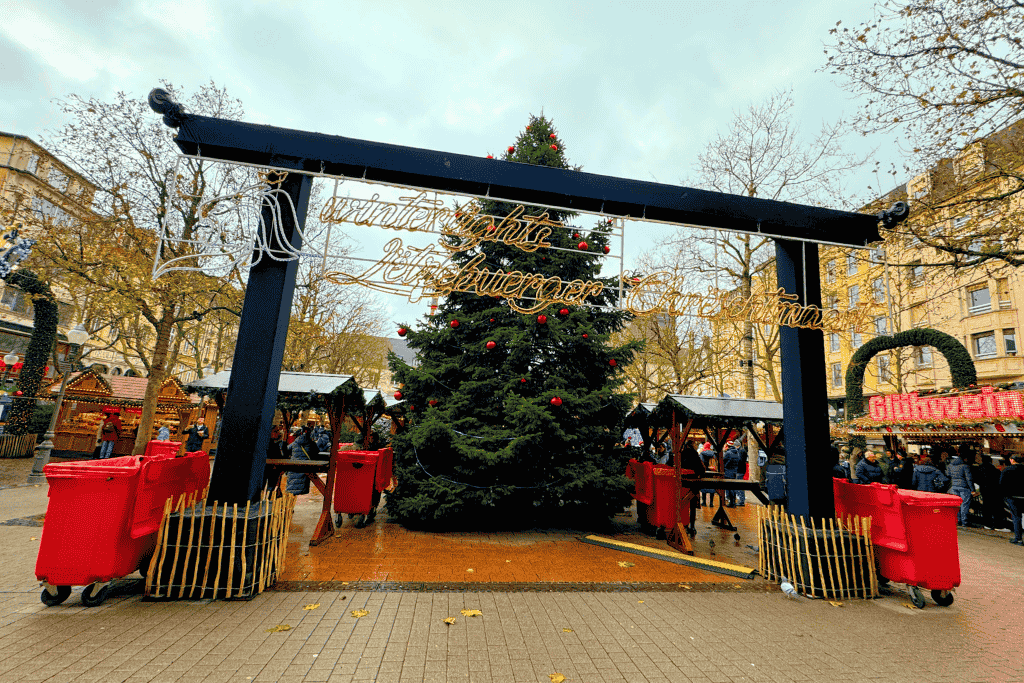 Kate walks through the entrance of the “Winterlights Lëtzebuerger Chrëschtmaart” in Luxembourg City, where a towering Christmas tree decorated with red ornaments stands at the center of the festive market.