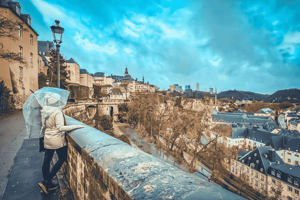 Kate stands with a clear umbrella overlooking the scenic cliffs and historic architecture of Luxembourg City, taking in the panoramic view of the old quarters and fortress walls.