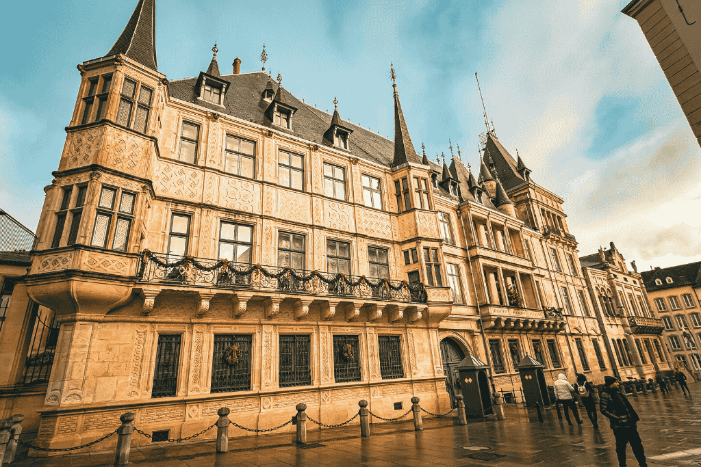 The Grand Ducal Palace in Luxembourg City stands with its ornate facade and pointed towers, glowing warmly in the late afternoon light as visitors pass by.