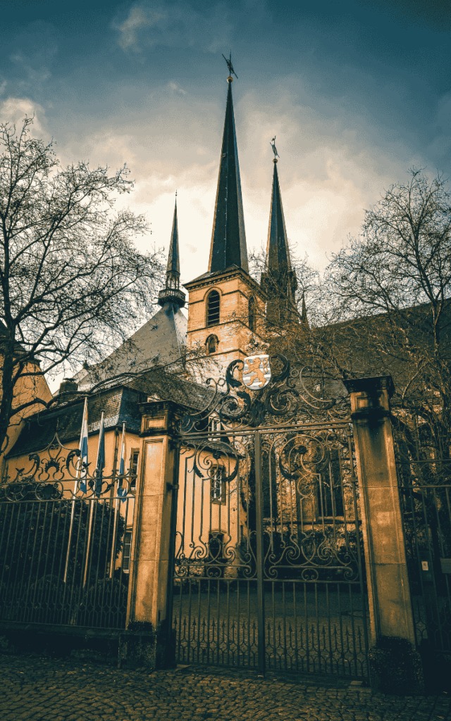The spires of Notre-Dame Cathedral in Luxembourg rise above ornate wrought-iron gates, framed by bare winter trees against a moody sky.