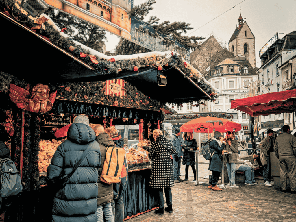 Visitors browse a festive food stall and gather under red umbrellas at the Basel Christmas market.