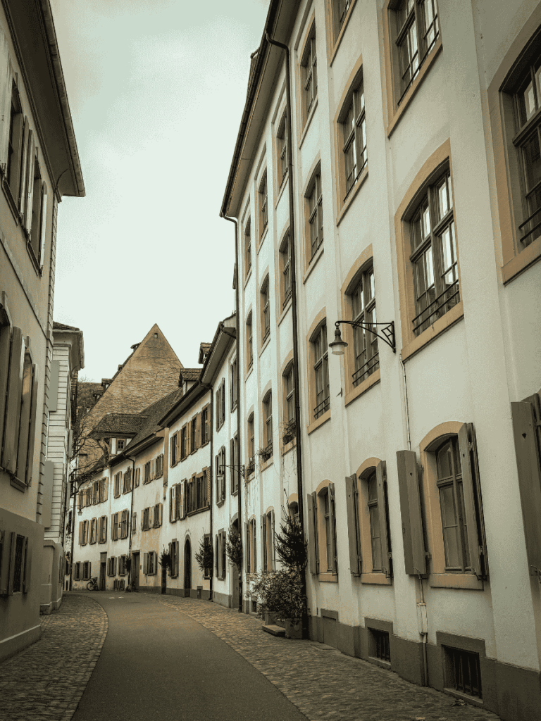 A quiet cobblestone street in Basel is lined with pastel buildings and shuttered windows.