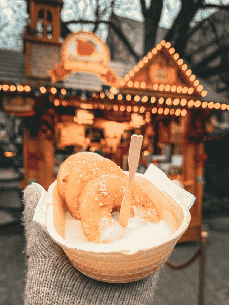 A gloved hand holds a tray of sugared pastries with cream in front of a glowing market stall.