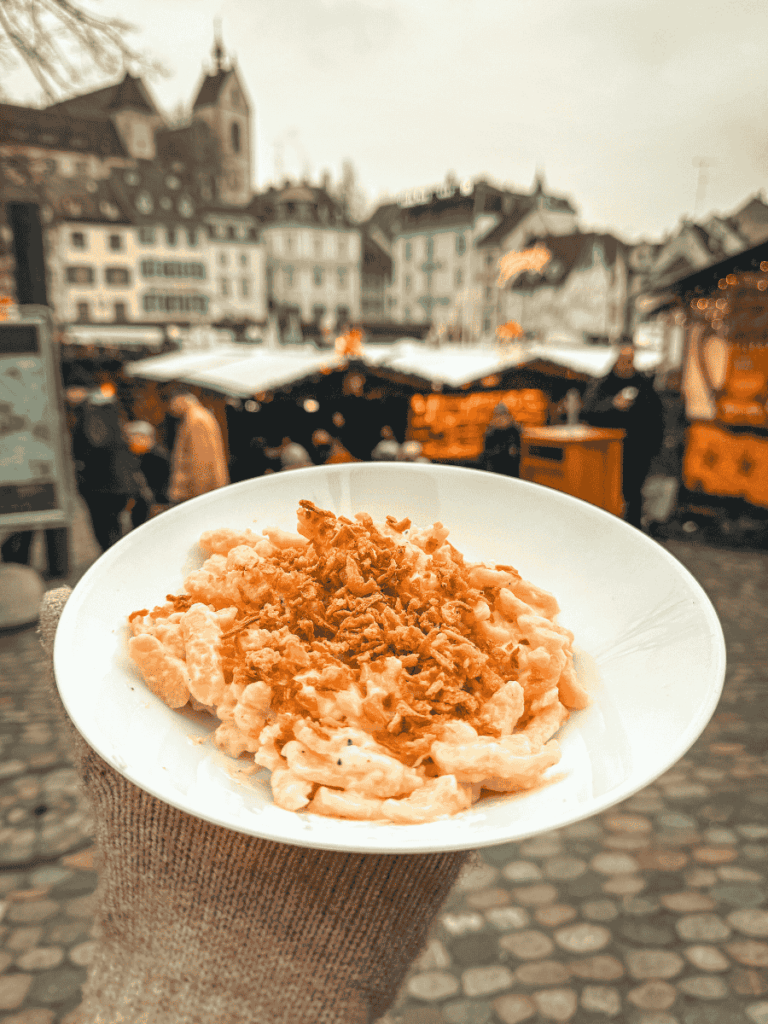 A gloved hand holds a bowl of cheesy spaetzle topped with crispy onions at the Basel Christmas market.