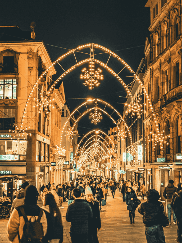 Crowds stroll beneath glowing arches of Christmas lights along a busy shopping street in Basel.