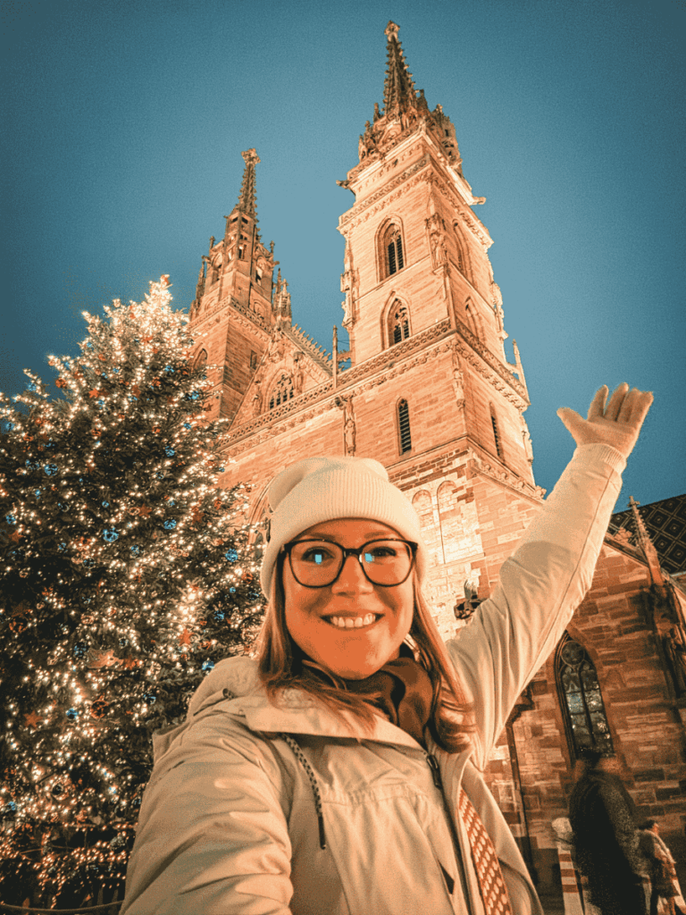 Kate smiles and gestures excitedly in front of Basel Minster and a glowing Christmas tree.