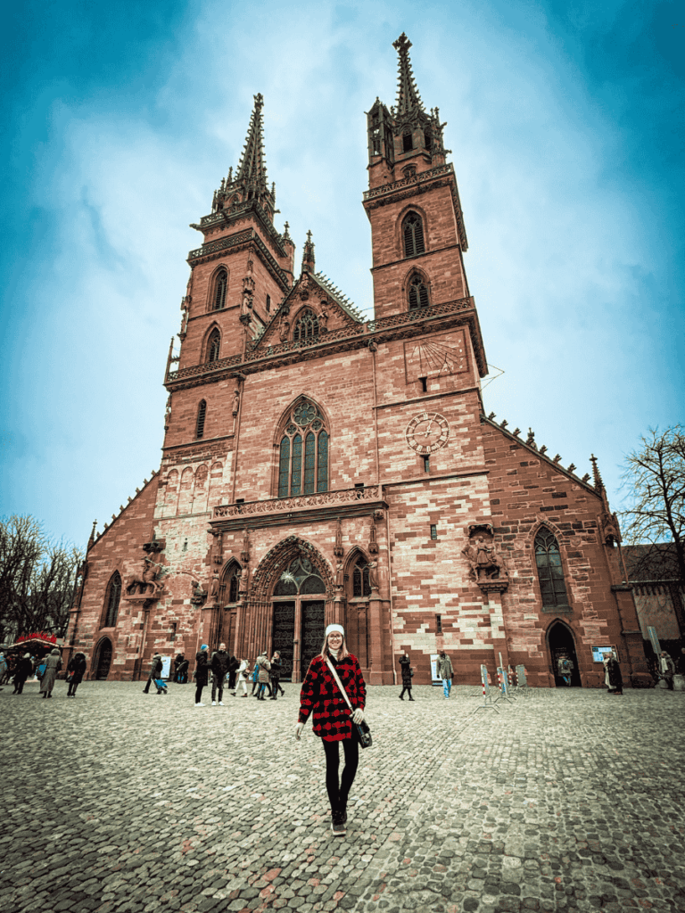 Kate stands smiling in front of the grand Basel Minster on a cobblestone square.