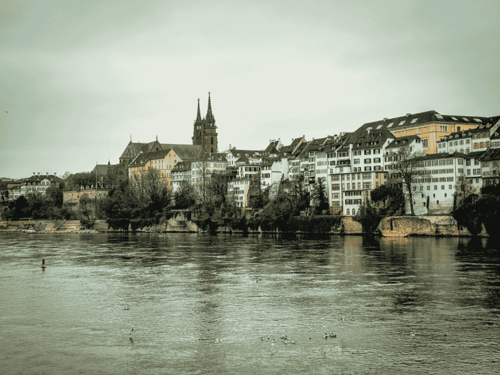 Colorful houses and the twin towers of Basel Minster overlook the Rhine River on a cloudy day.