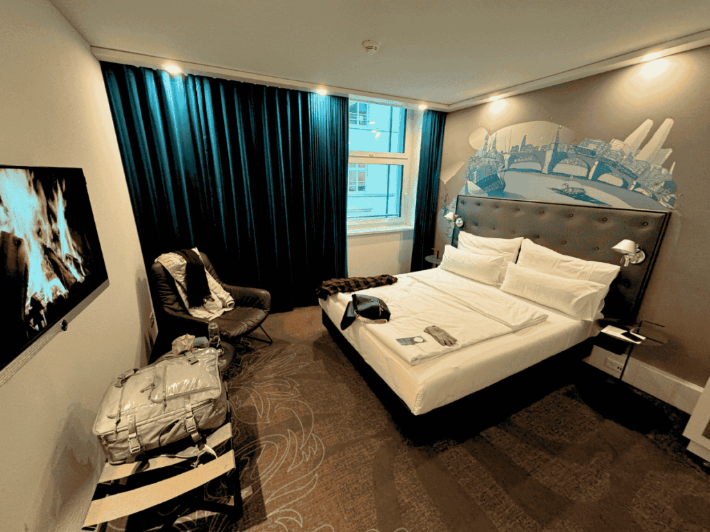 A cozy hotel room in Basel features a neatly made bed, cityscape wall art, and a faux fireplace on the TV.