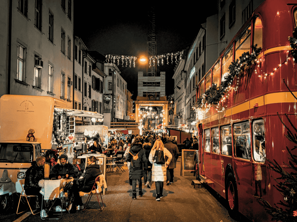 Visitors browse a festive food stall and gather under red umbrellas at the Basel Christmas market. You said: ChatGPT said: The Basel Christmas market glows at night with twinkling lights, decorated stalls, and festive crowds. You said: ChatGPT said: People enjoy food and drinks at festive street stalls beside a decorated double-decker bus in Basel at night.
