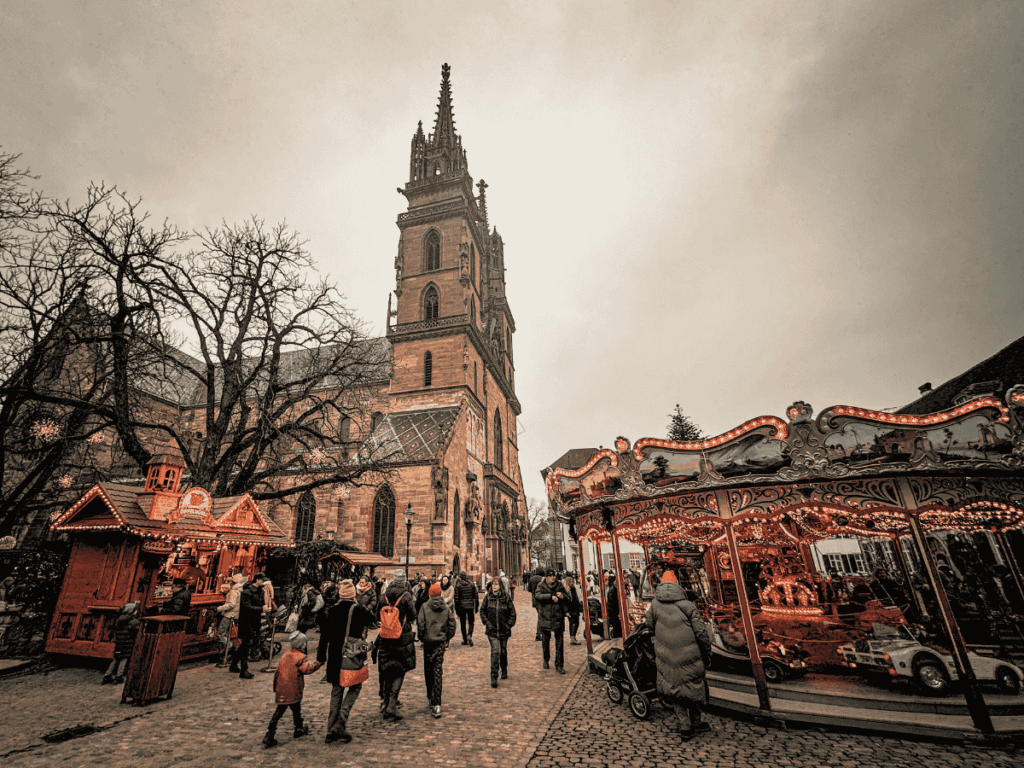 Families stroll past a glowing carousel and festive stalls beside Basel Minster at Christmas.