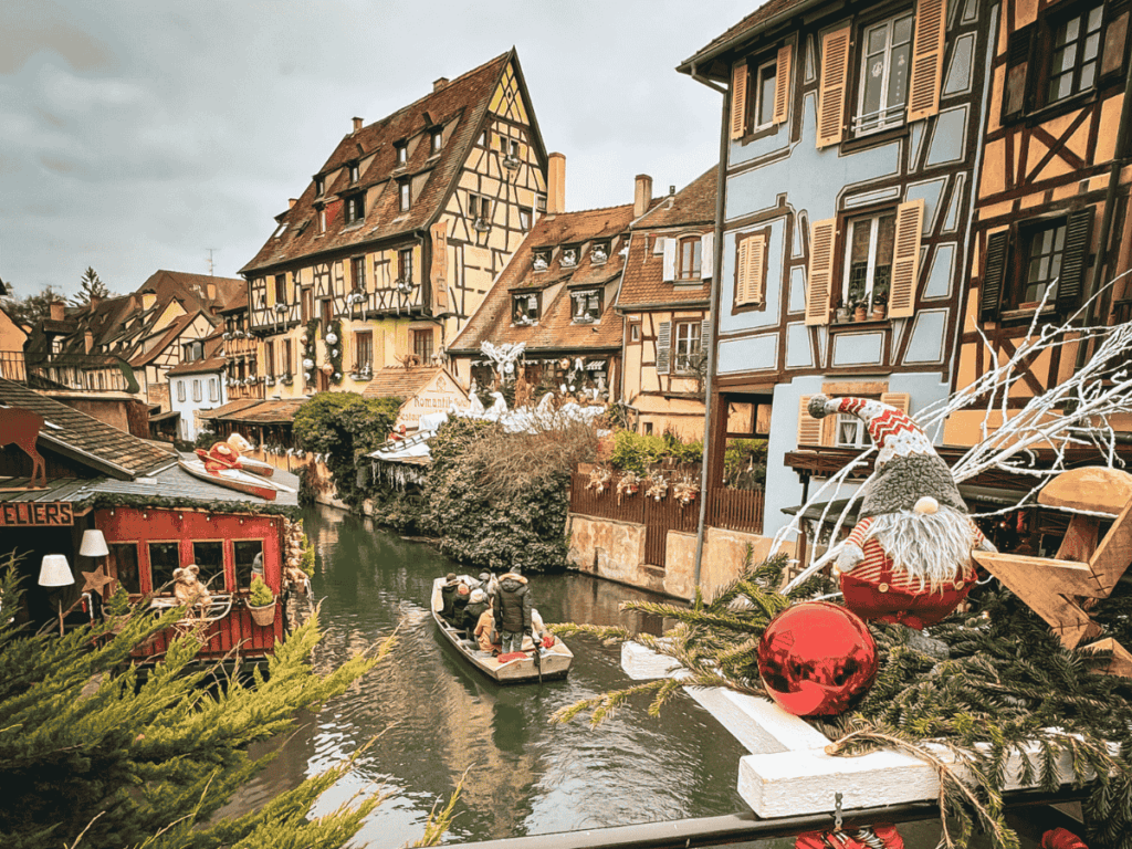 A decorated canal in Colmar is lined with colorful half-timbered houses as a small boat glides past Christmas ornaments and festive displays.