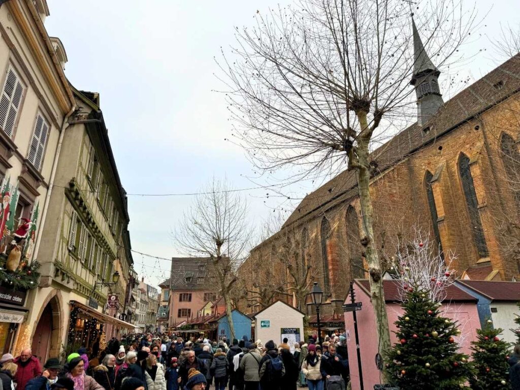 A bustling crowd explores the festive Christmas market stalls along a historic Dominican Church in Colmar.