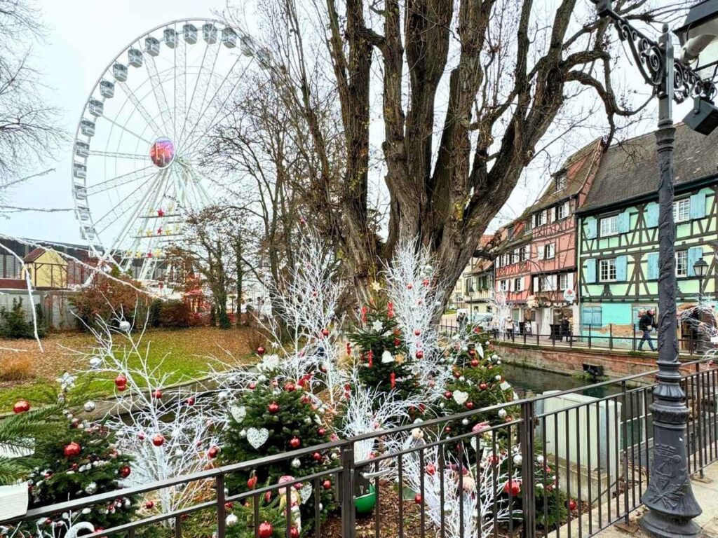 A festive scene in Colmar shows decorated Christmas trees by the canal with a Ferris wheel in the background.