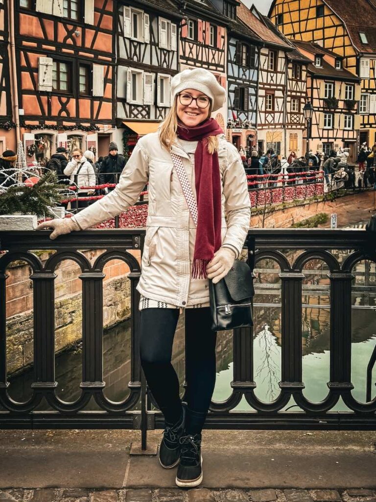 Kate smiles by a canal railing with colorful half-timbered houses behind her in Colmar.