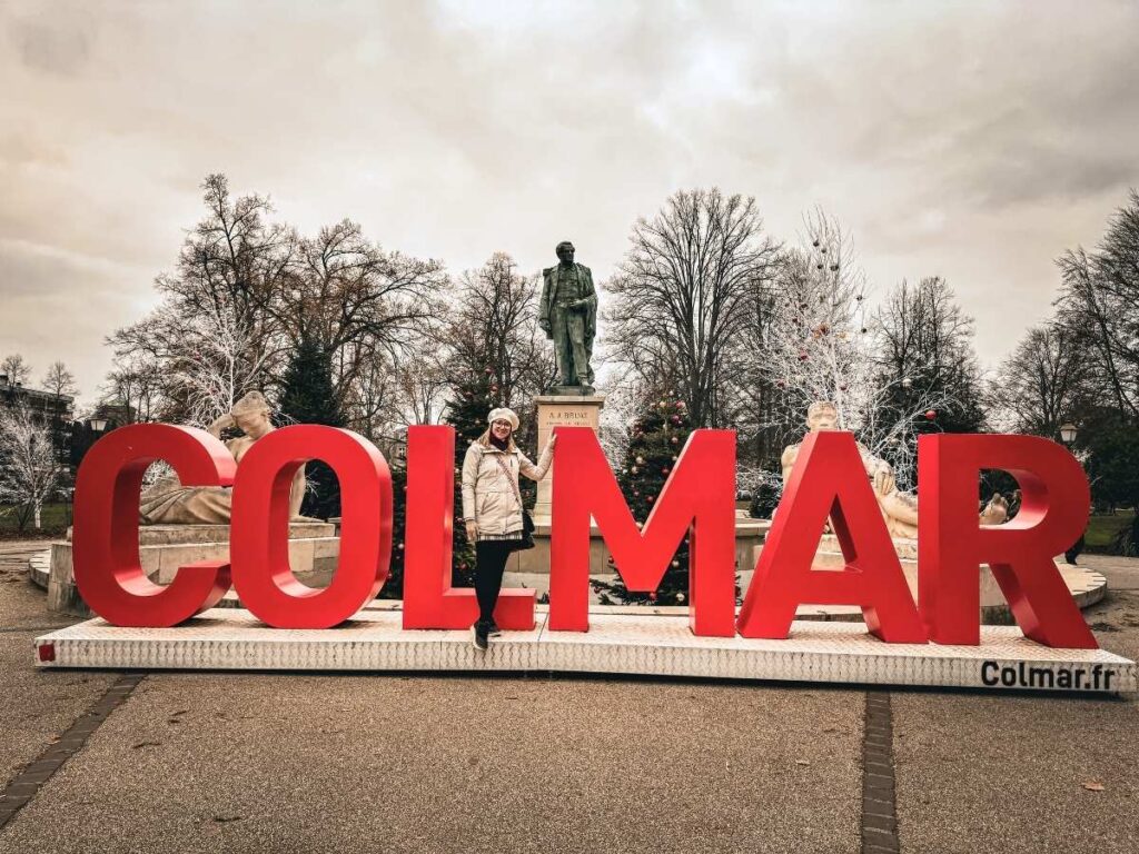 Kate poses inside the large red "Colmar" sign decorated for Christmas in a town square.