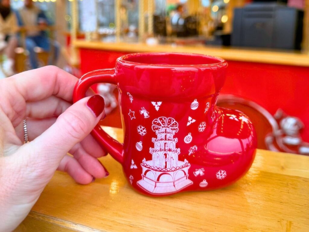 A red boot-shaped Christmas mug with festive designs is held up at a Colmar market stall.