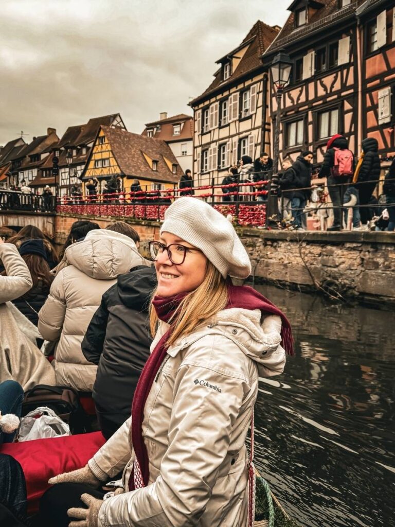 Kate smiles while sitting on a canal boat surrounded by festive half-timbered buildings in Colmar.