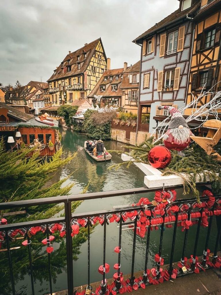 A canal boat passes colorful half-timbered houses in Colmar, viewed from a bridge decorated with red locks and Christmas ornaments.