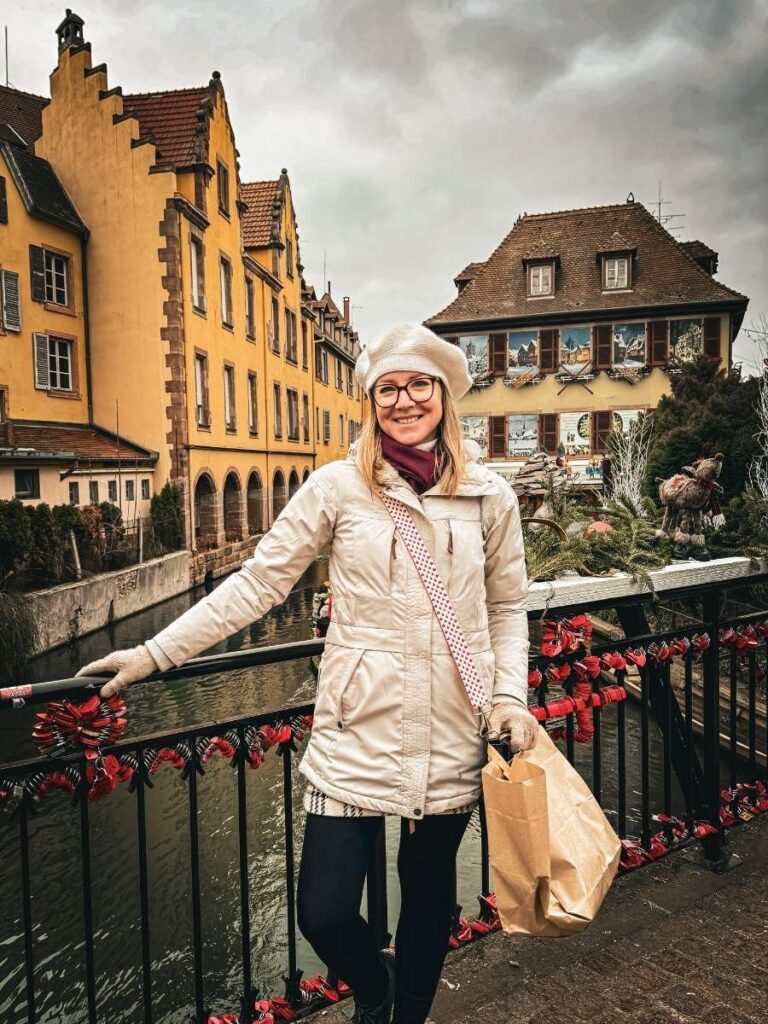 Kate poses on the decorated bridge in Colmar with festive buildings and the canal behind her.