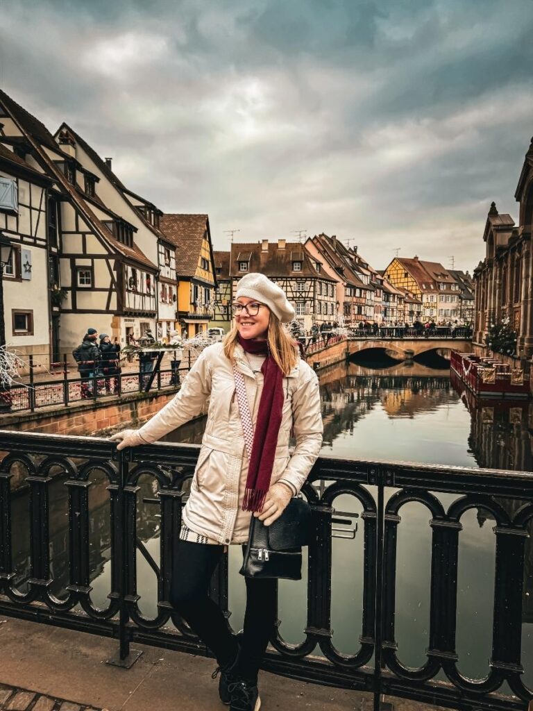 Kate poses on a bridge in Colmar with half-timbered houses and a stone canal bridge in the background.