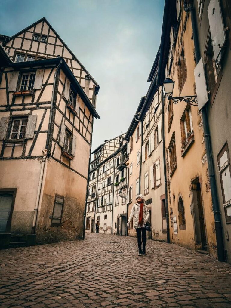 Kate walks along a quiet cobblestone street lined with historic half-timbered houses in Colmar.