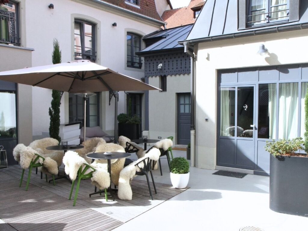 A cozy courtyard patio is set with tables, fur-covered chairs, and a large umbrella outside a hotel in Colmar.