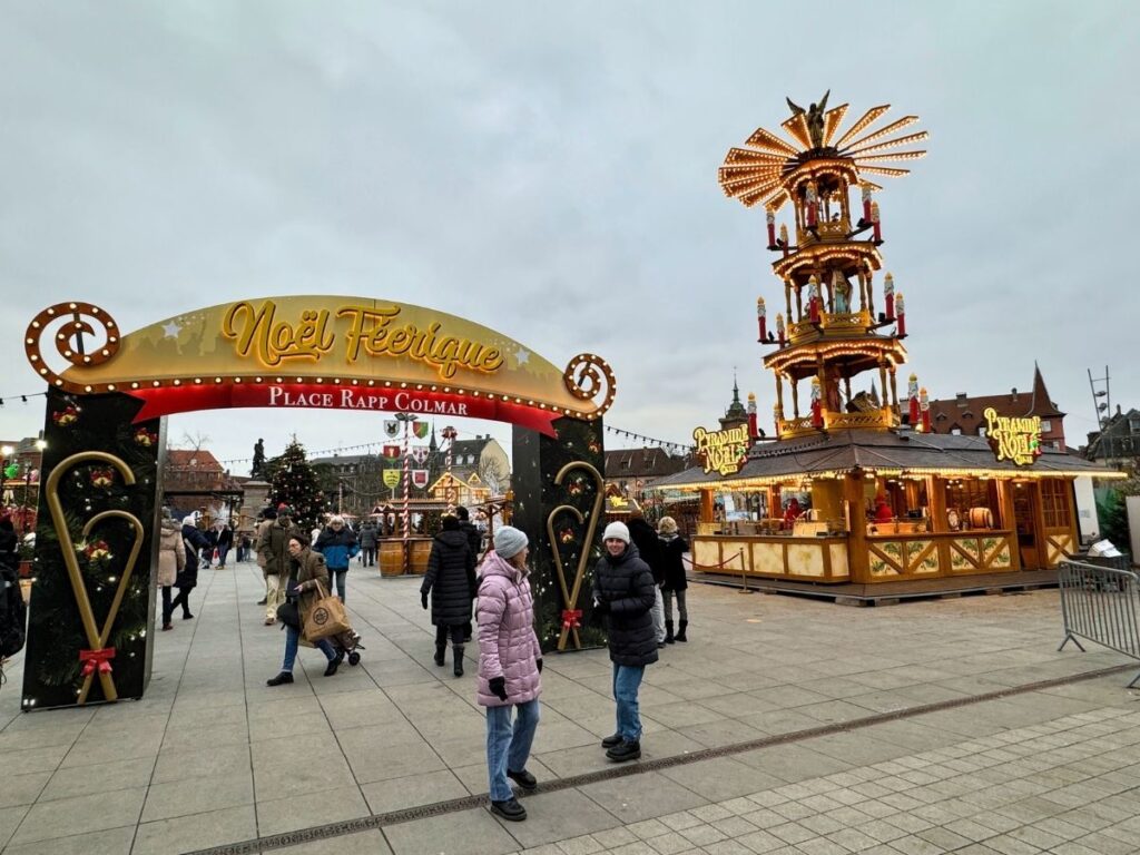 People stroll through Colmar’s Place Rapp Christmas market under the “Noël Féérique” archway beside a tall illuminated wooden pyramid.