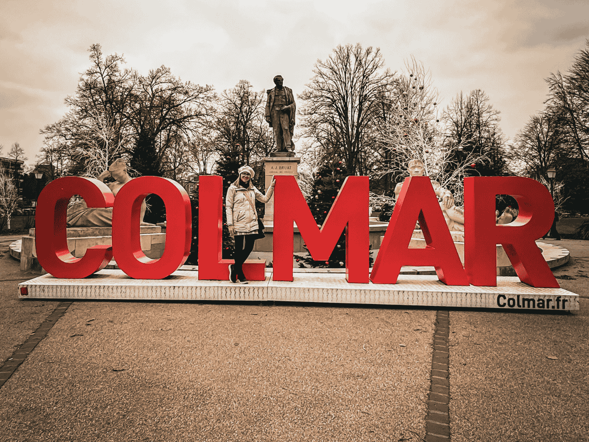 Kate stands inside giant red “Colmar” letters in a festive square decorated for Christmas.