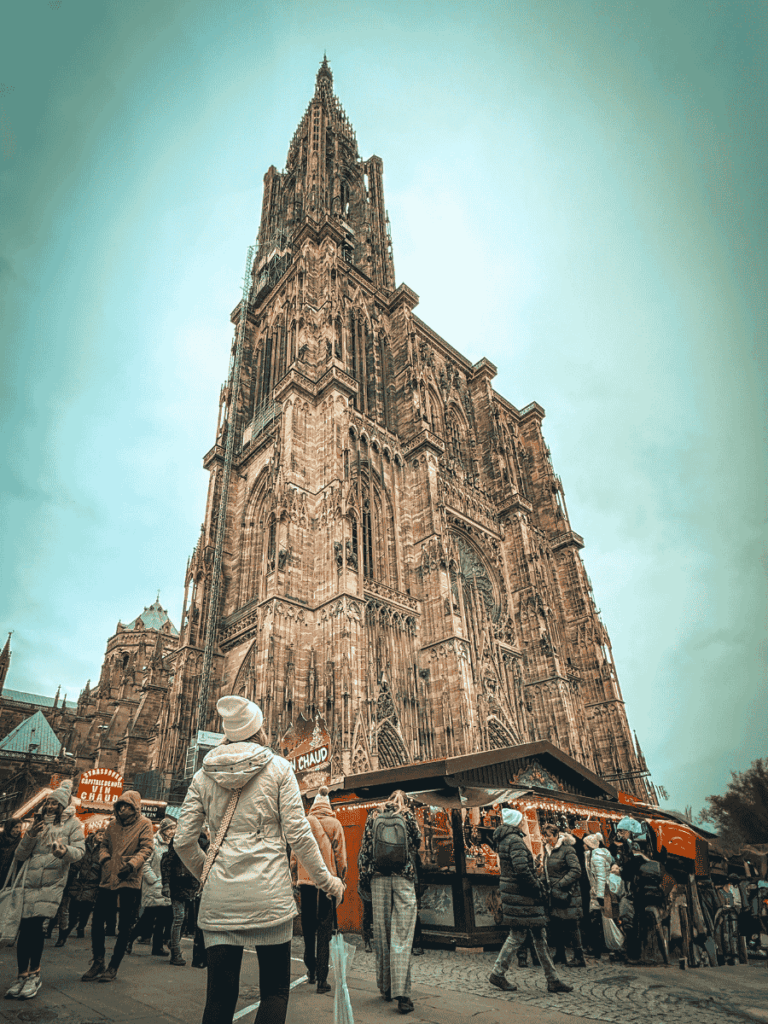 Kate, wearing a white coat and hat, stands in front of the immense Strasbourg Cathedral, gazing upward while holding an umbrella among the Christmas market crowd.